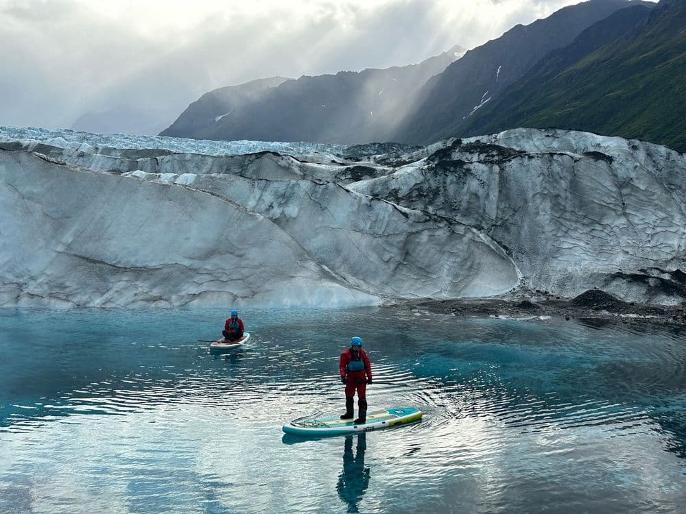 Two paddleboarders on turquoise meltwater below a towering glacier face, sunbeams breaking through cloud