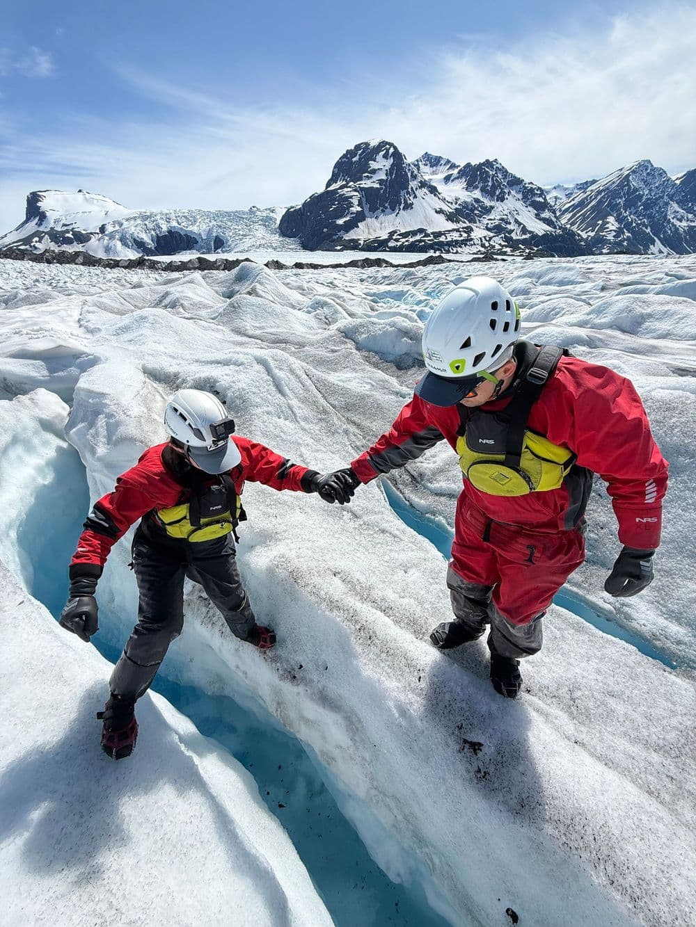 Two guides in red drysuits and climbing helmets steadying each other across a crevasse on a glacier
