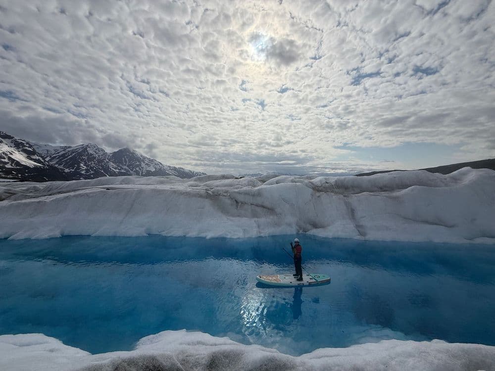 A lone paddleboarder crossing a brilliant blue meltwater pool on top of a glacier, beneath a mackerel sky